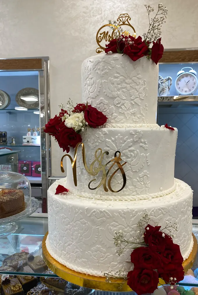 Gâteau de mariage à trois étages en dentelle blanche, roses rouges et gypsophile, monogramme et cavalier dorés, sur commande.
