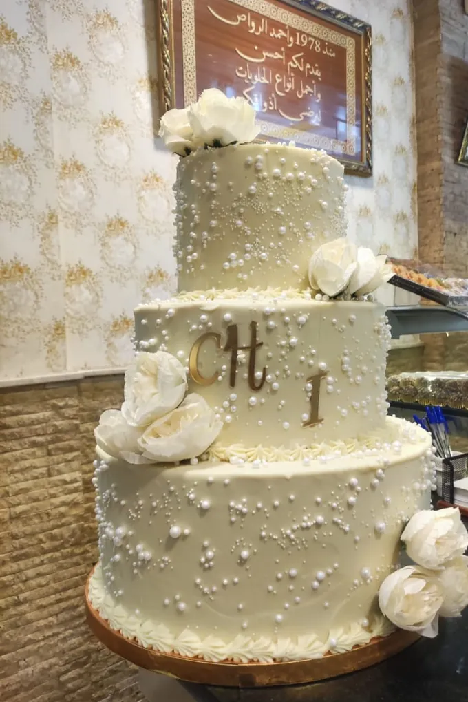 Gâteau de mariage à trois étages entièrement perlé de blanc, fleurs de sucre ivoire et monogramme doré, sur commande.
