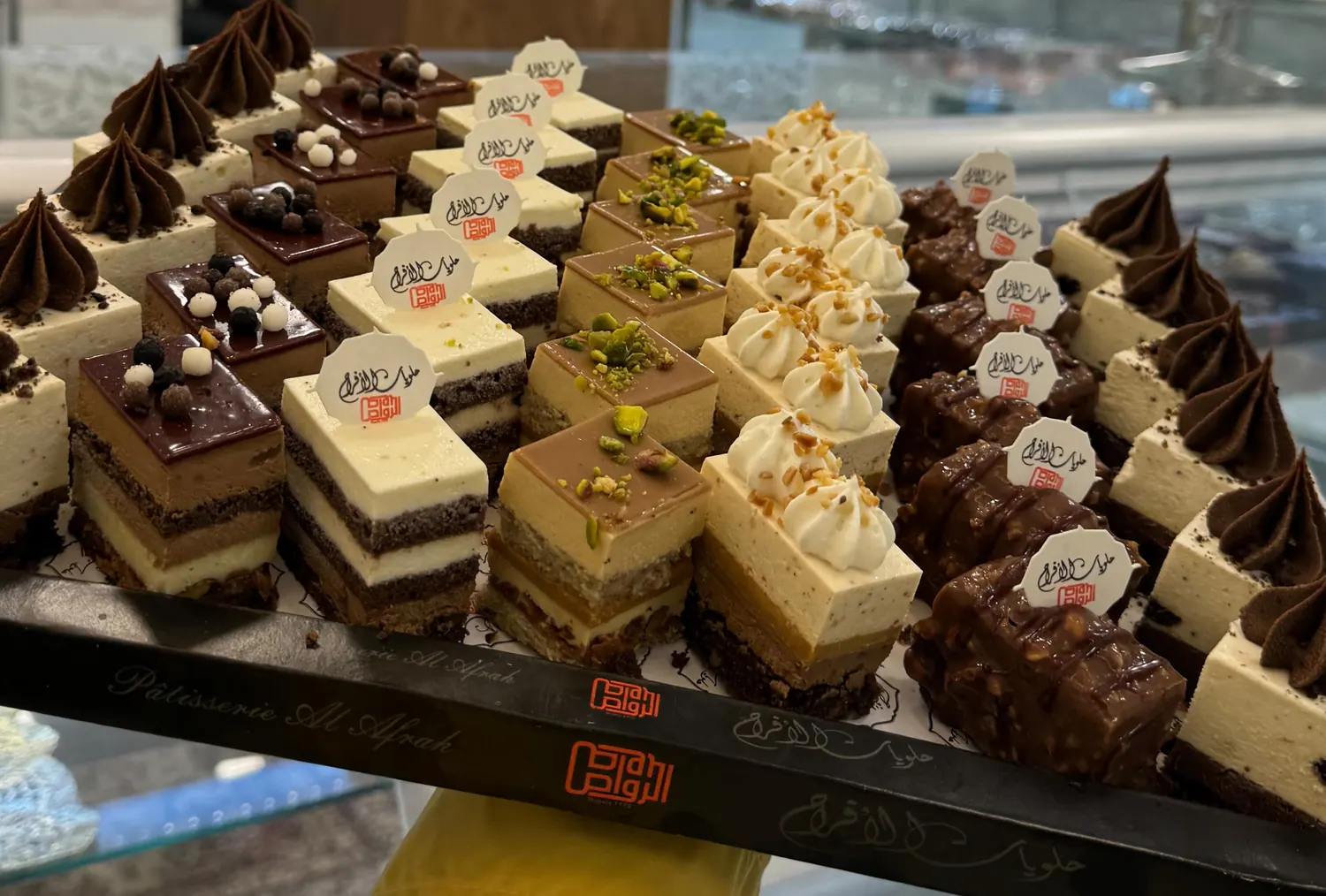 Un plateau de nos gâteaux individuels (bavarois au chocolat, à la pistache et aux fruits secs), présenté au comptoir, la mention « Pâtisserie Al Afrah » gravée sur le bord du plateau.